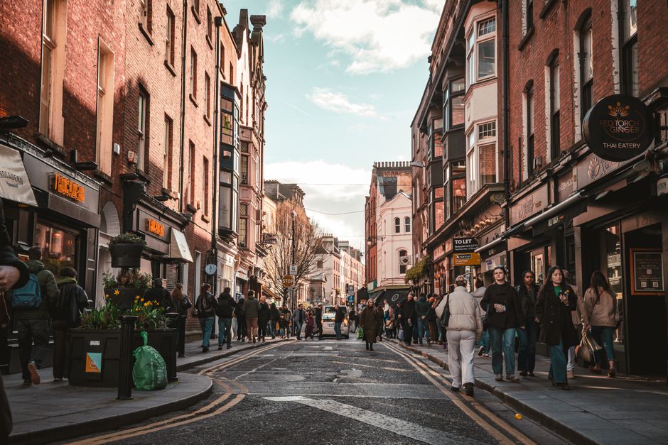 A busy street scene in Dublin's Temple Bar district showing pedestrians walking along the pavement and crossing the road, with a variety of shops and restaurants on both sides. Storefronts display sig