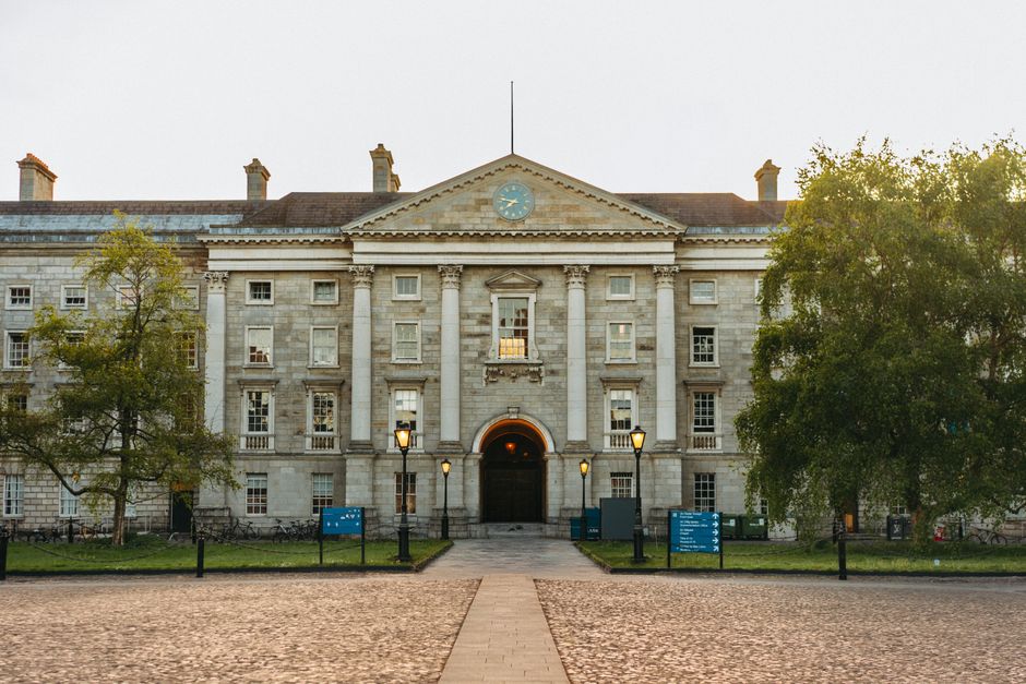 A symmetrical, neoclassical-style building with a stone facade, featuring a central entrance with a rounded arch doorway and illuminated street lamps lining the paved pathway leading to it. The buildi