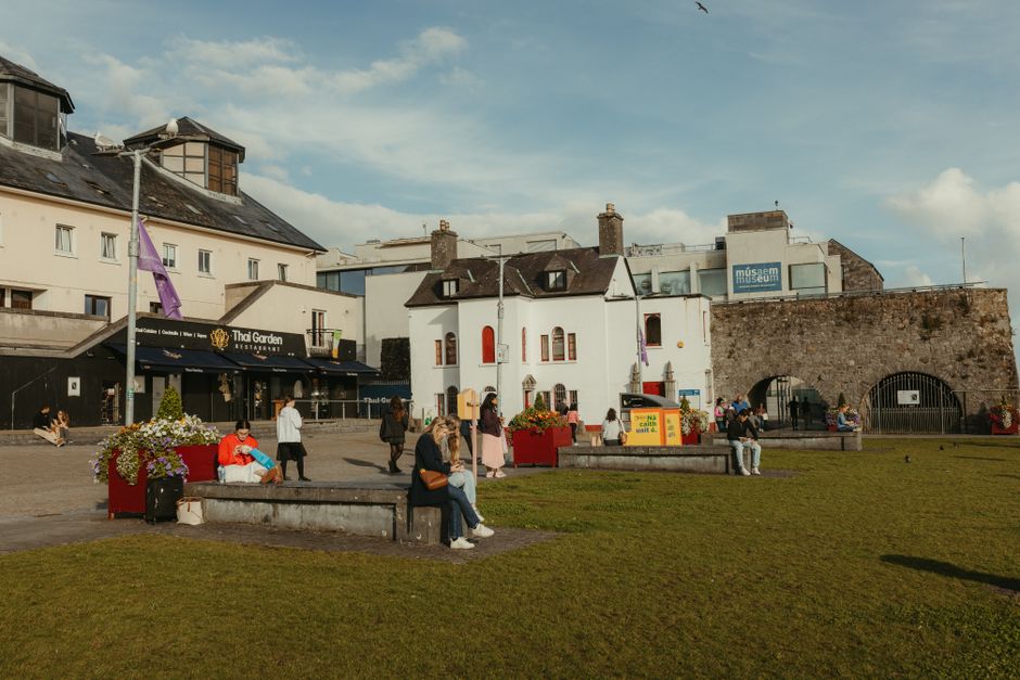 A symmetrical, neoclassical-style building with a stone facade, featuring a central entrance with a rounded arch doorway and illuminated street lamps lining the paved pathway leading to it. The buildi